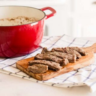 Red dutch oven with round steak with sliced round steak on wooden cutting board.