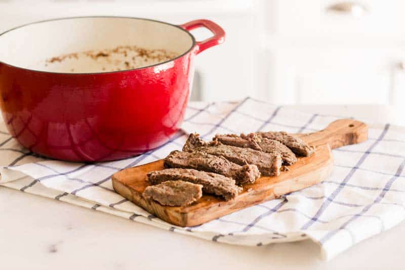 Red dutch oven with round steak with sliced round steak on wooden cutting board.