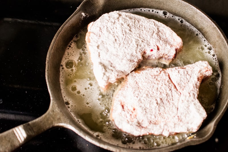 Batter coated pork chops frying in a cast iron skillet on the stovetop.