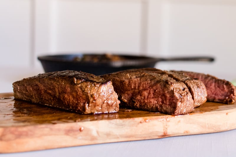 Medium rare venison steak, sliced on a cutting board with a cast iron skillet.