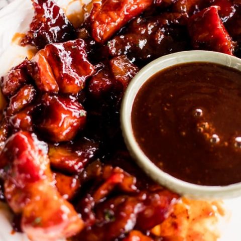 Pork belly burnt ends on white plate with a ramekin of bbq sauce.