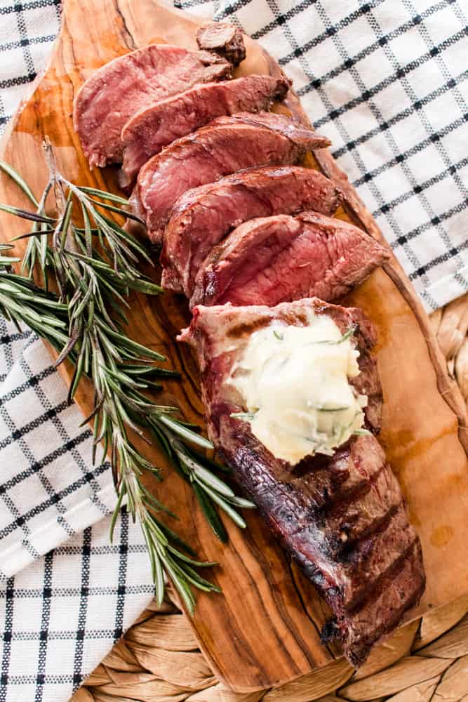 Elk tenderloin cooked and sliced on a white checkered tea towel with rosemary springs.