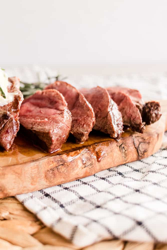 Elk tenderloin cooked and sliced on a white checkered tea towel with rosemary springs.