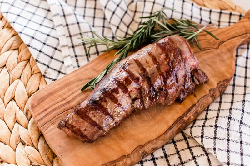 Homemade elk tenderloin seared on a grill pan with rosemary sticks on a wicker mat.