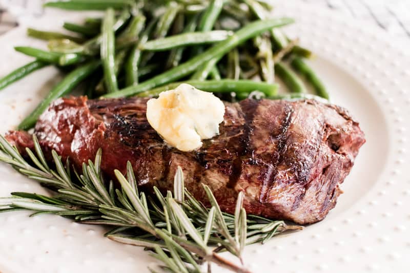 Elk backstrap dinner recipe on a white plate with a dallop of rosemary garlic butter and green beans.