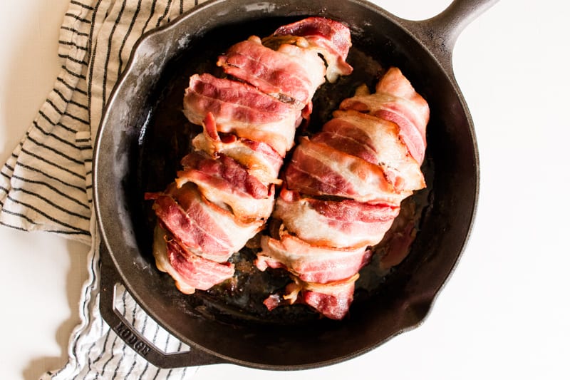 Bacon wrapped stuffed venison backstrap in a cast iron skillet on a stripped tea towel.