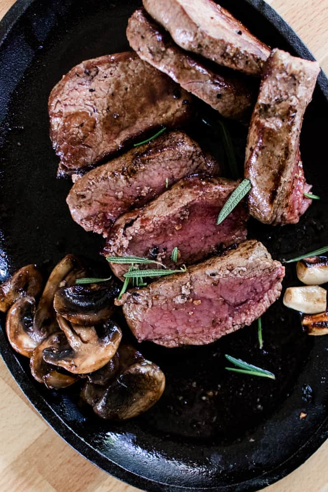 Butterfly elk steak medallions sliced up on a black plate with mushrooms and garnished with fresh rosemary.