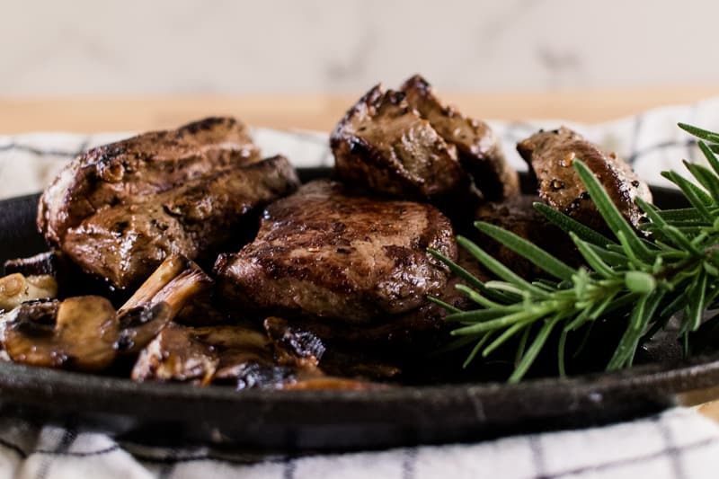 Elk steak dripping in butter on a oval cast iron serving dish with a spring of rosemary and sautéed mushrooms with it.