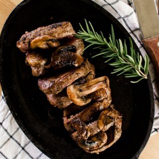 Elk tenderloin steak on a sizzle platter with a spring of rosemary, fork, knife on top of a wooden table.