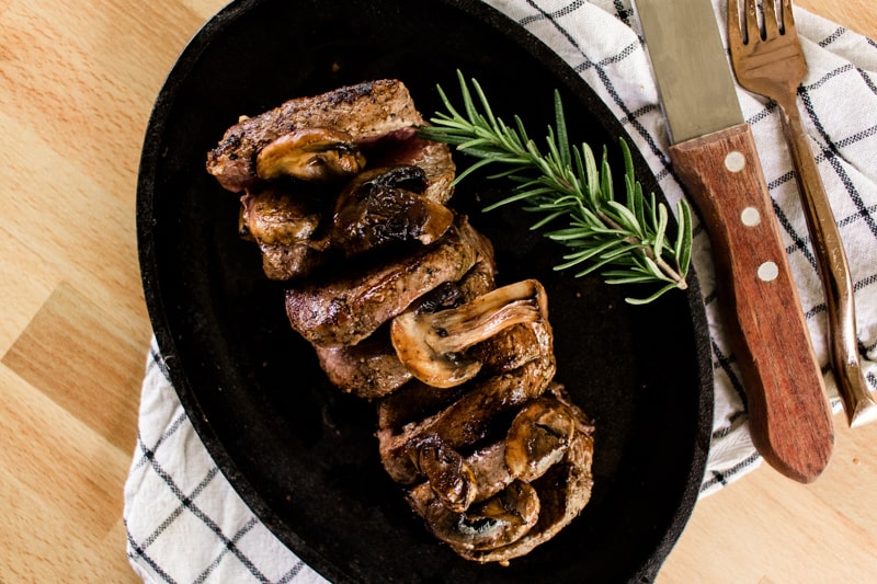 Elk tenderloin steak on a sizzle platter with a spring of rosemary, fork, knife on top of a wooden table.