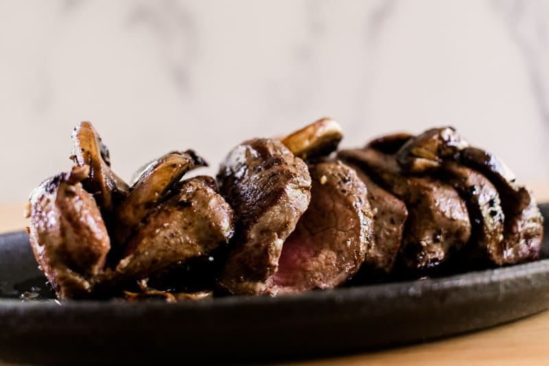 Elk steak cut up like a tenderloin topped with savory mushrooms and garlic on a cast iron serving plate.