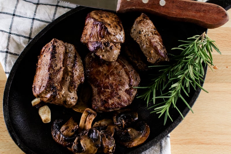 Elk steaks with a steak knife, rosemary, sautéed butter and garlic cloves on a wooden tabletop.