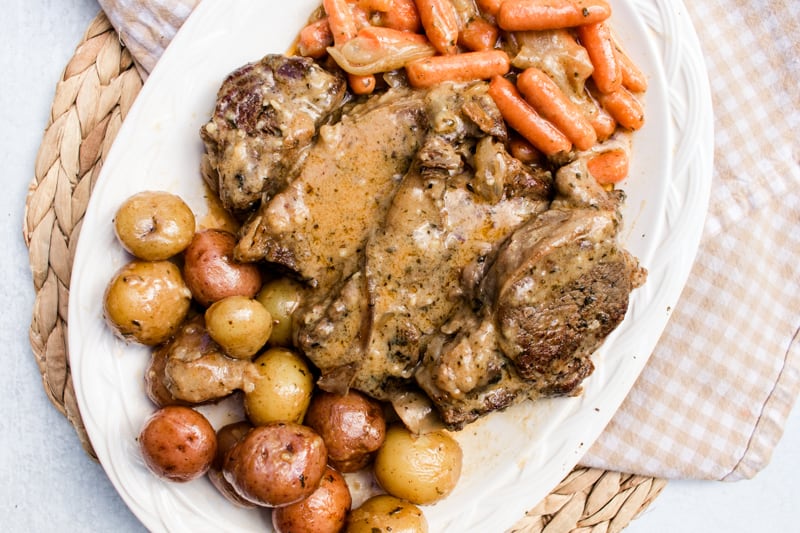 Chuck roast smothered in gravy on a large white plate with cooked potatoes and carrots.