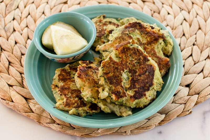 Fried zucchini fritters on a teal plate with sliced lemons.