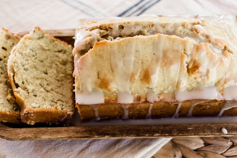Tender zucchini bread sliced on a wooden bread tray with a white glaze drizzled over the top.