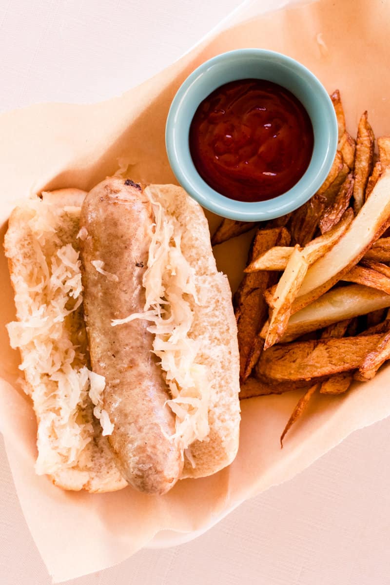 Bratwurst cooked on the stove placed in a bun with sauerkraut, served with ketchup and homemade french fries. 