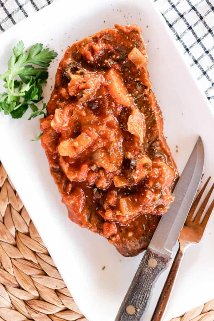 Carne pizzaiola steak in tomato sauce served on a white dish on a table top. 