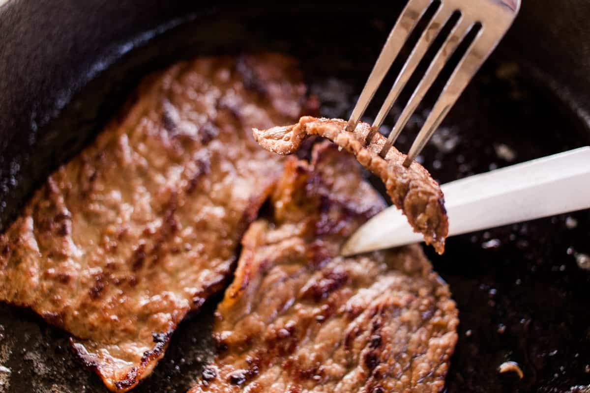 A tender slice of Marinated Bottom Round Steak on a fork with the steak and knife in the background.