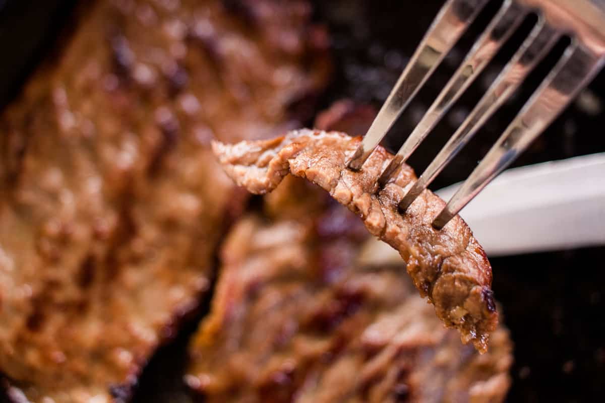 A tender slice of Marinated Bottom Round Steak on a fork with the steak and knife in the background.