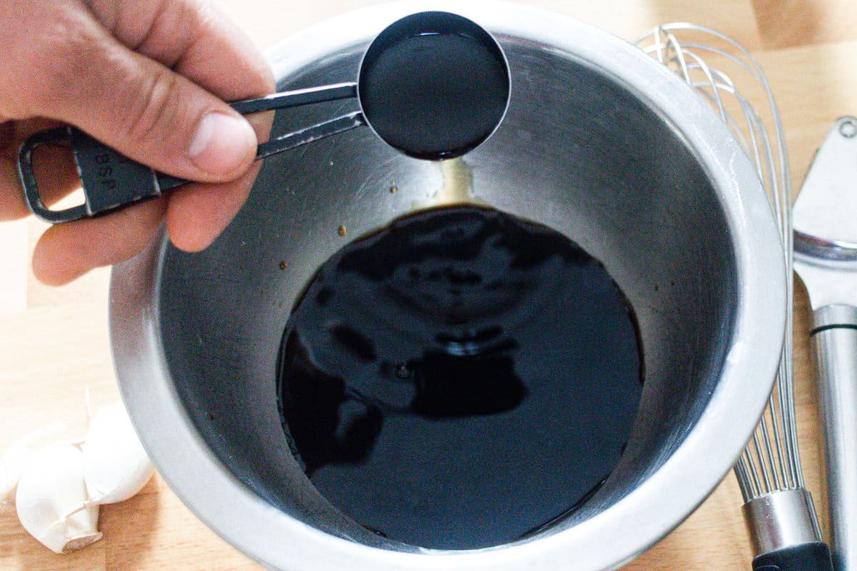 A measuring spoon full of soy sauce being added to metal bowl with marinade ingredients, surrounded by cloves of garlic, a whisk, and a garlic press.