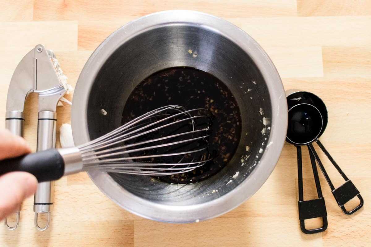 A metal bowl with marinade ingredients being whisked together, surrounded by a garlic press and measuring spoons.