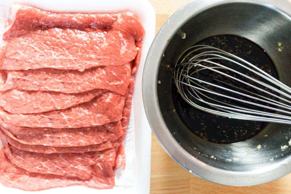 A tray of bottom round steak next to a metal bowl and whisk with and dark marinating sauce.