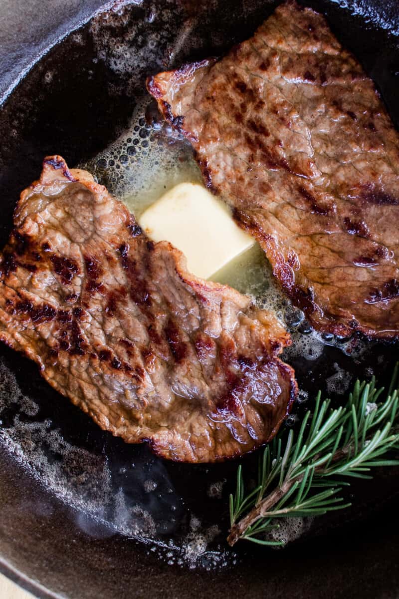 Nicely browned bottom round steaks cooking in a cast iron skillet with butter and rosemary.