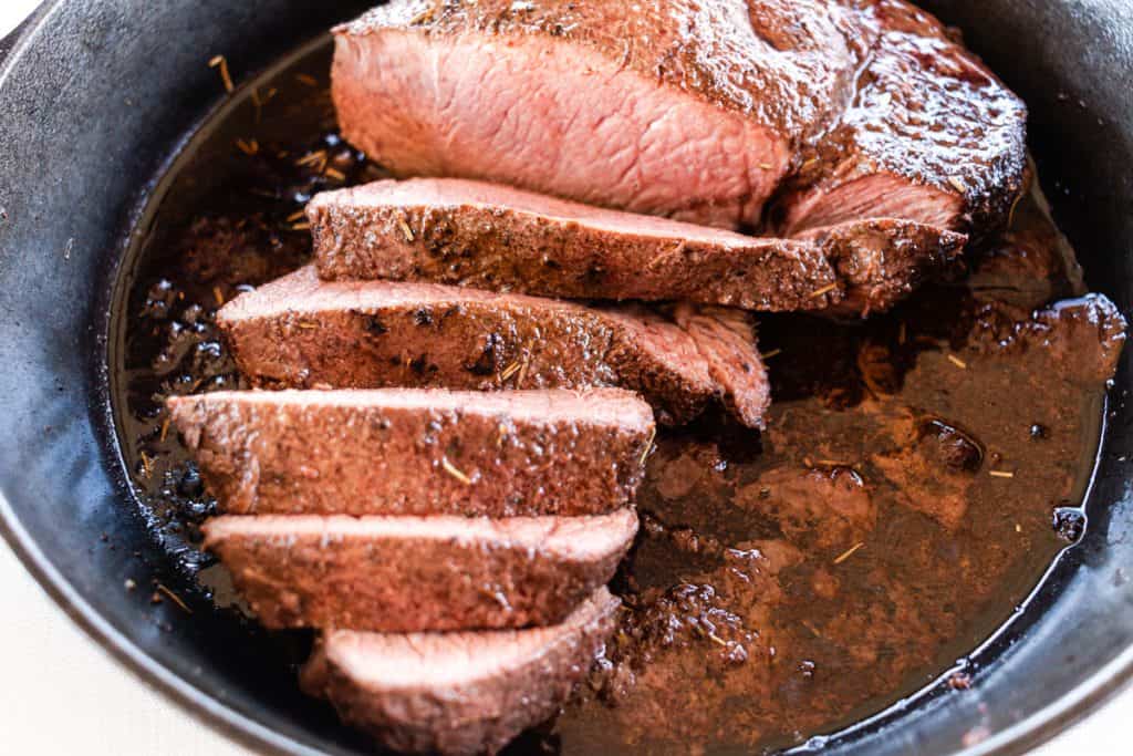 Slices of perfectly cooked sirloin tip roast sitting in its juices in a cast iron skillet.