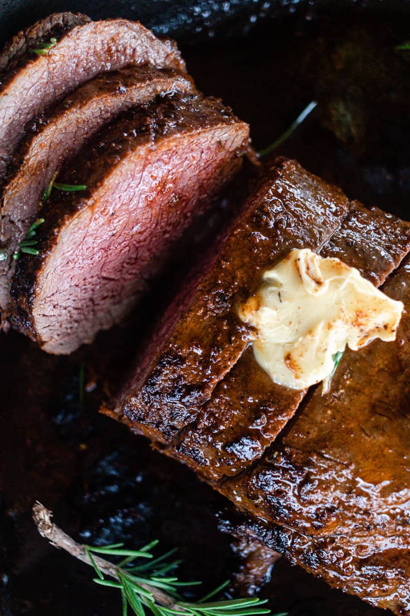 Slices of mouth-wateringly delicious deer tenderloin with fresh rosemary sprigs and a dollop of butter on a black cast iron skillet.