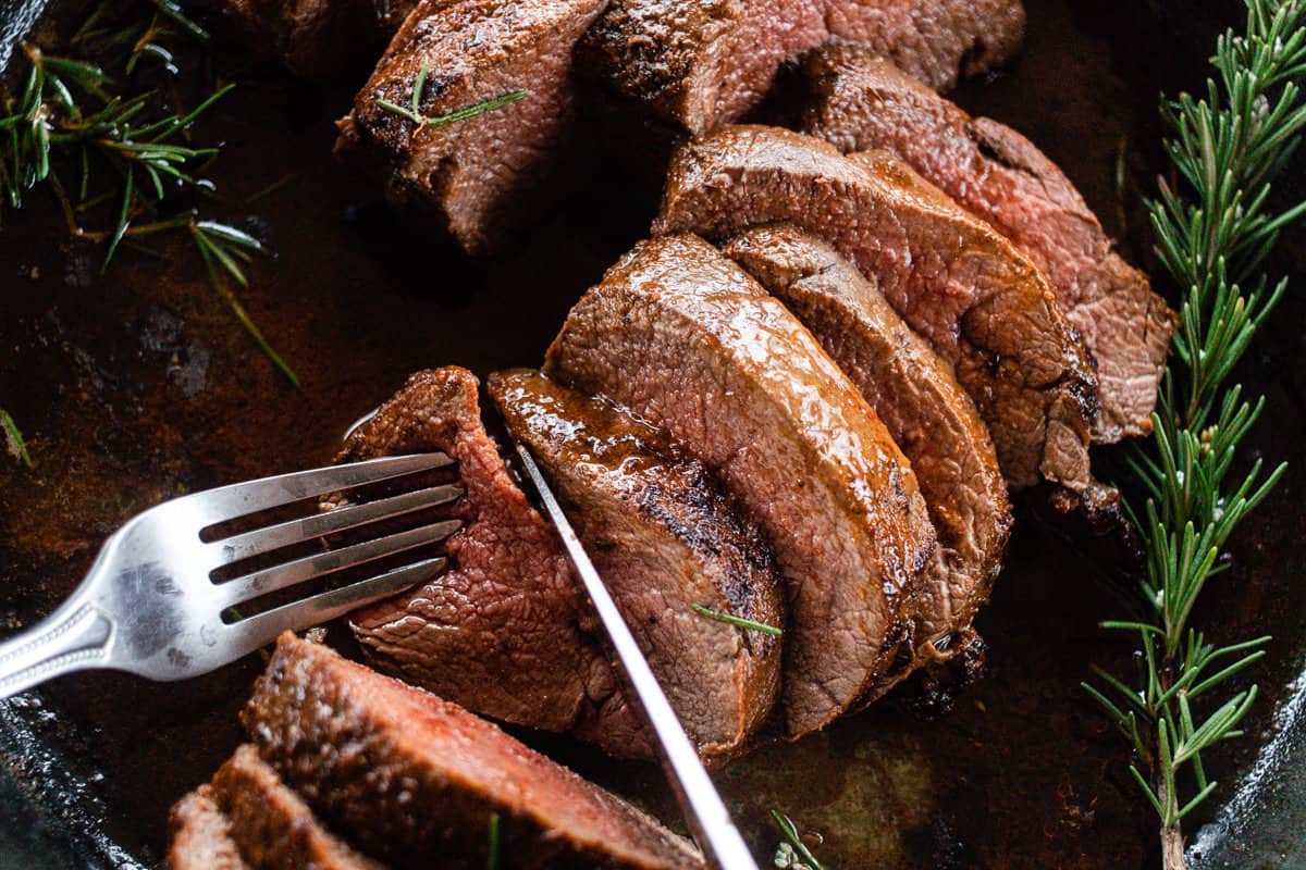 Slices of beautifully cooked deer tenderloin topped with fresh rosemary sprigs on a black cast iron skillet, with a fork and knife slicing into the meat in the foreground.