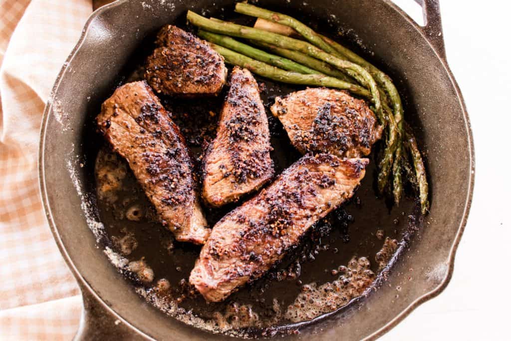 A cast iron pan with beautifully seared beef tri tip steak in a peppercorn rub with lightly cooked asparagus spears.