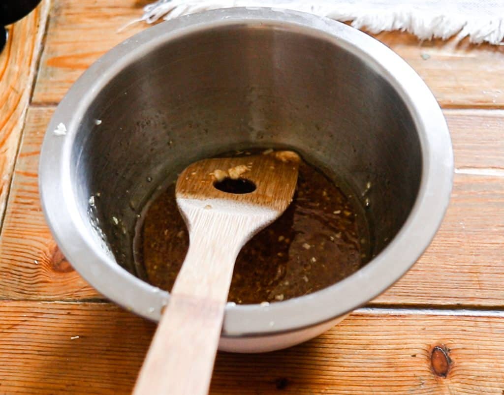 A metal bowl containing both a dark marinade liquid and a wooden spoon sitting on a wooden table.