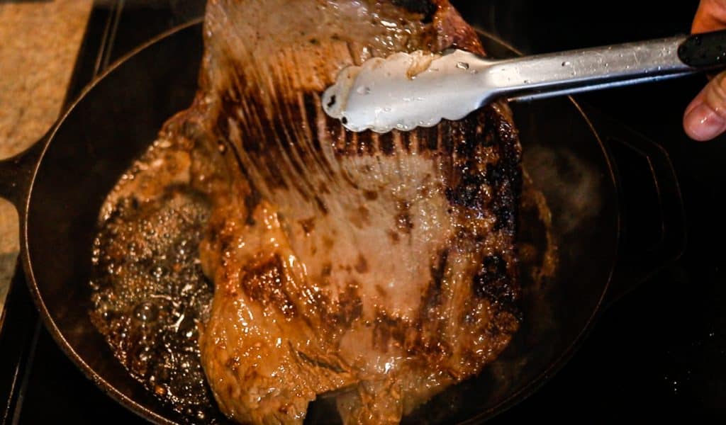 A flank steak in a black cast iron skillet being flipped with metal tongs.