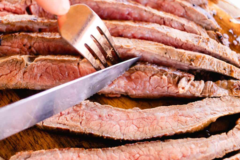 Slices of perfectly cooked flank steak being cut on a wooden cutting board.