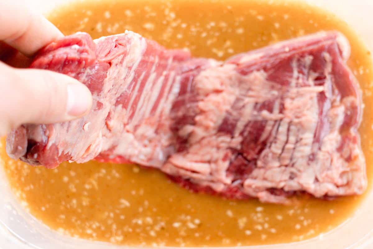 A skirt steak being lowered into an orange-colored marinade.