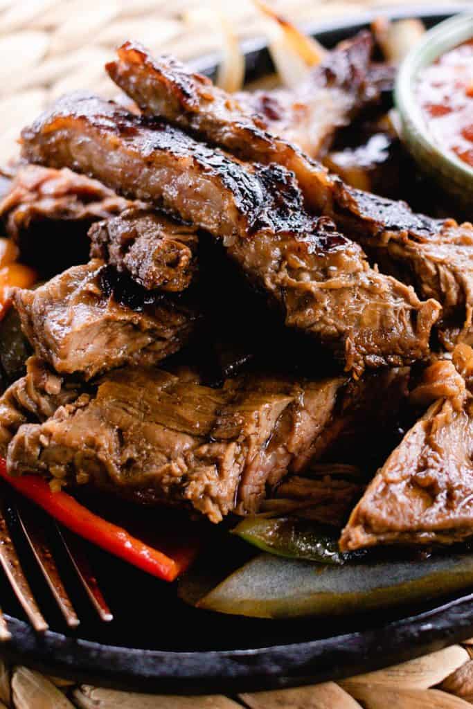 A black serving dish full of tender slices cast iron skirt steak fajita beef served over vegetables with a side of salsa just visible in the top right corner.