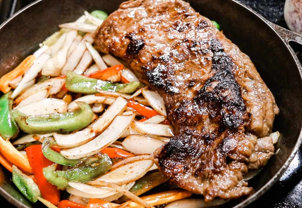 A black cast iron skillet containing white, red, and green vegetables beside a nicely browned skirt steaks.