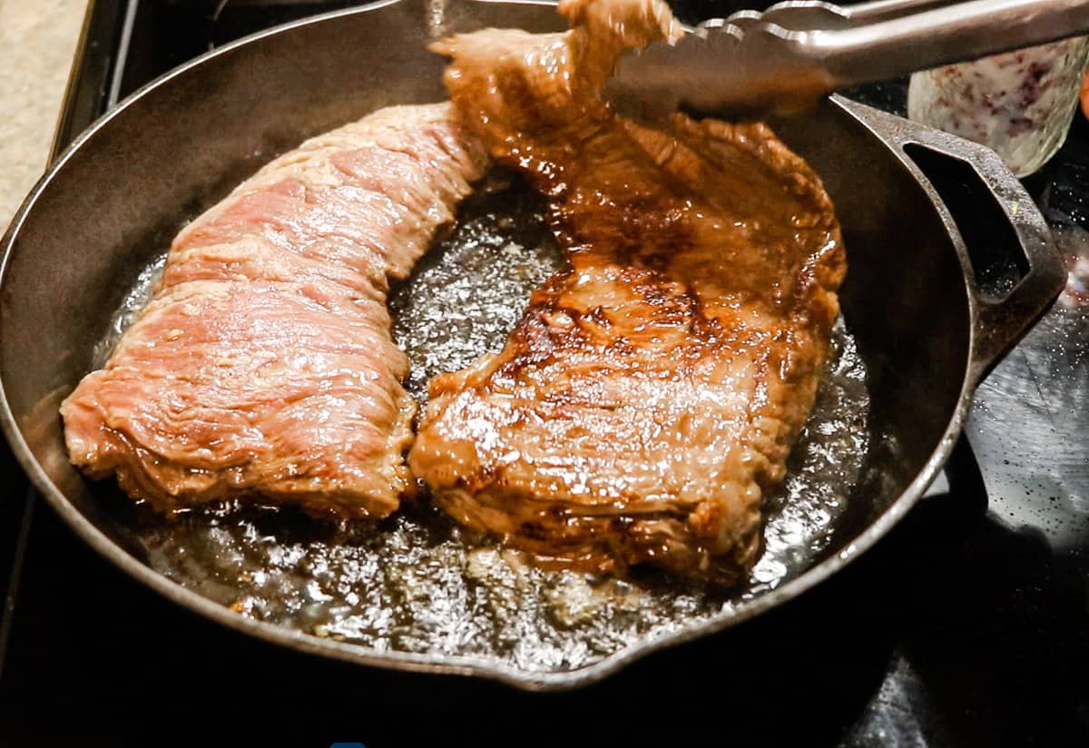 Two skirt steaks sizzling in a cast iron skillet — one has just been flipped with metal tongs.