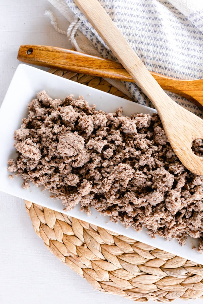 A white serving dish of boiled ground beef with wooden spoons and tea towels on a raffia trivet — ready for whatever delicious beefy meal you have planned. 