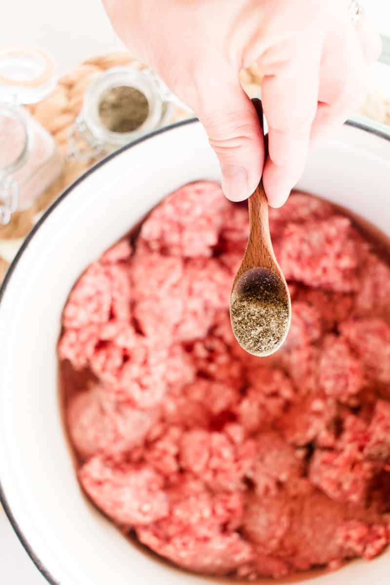 A dutch oven full of raw ground beef ready to be sprinkled with pepper by a hand holding a small wooden measuring spoon.
