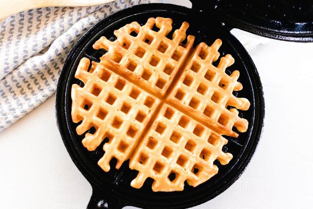 A golden waffle ready to be removed from a black, cast iron waffle iron, beside a blue and white tea towel.