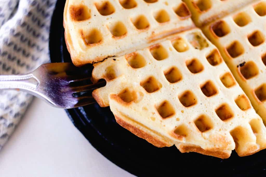 A fork lifting the edge of a golden waffle from its cast iron waffle iron, ready for serving.