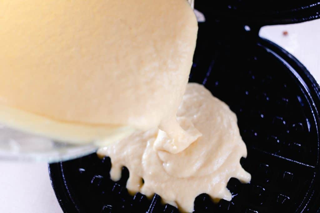 Waffle batter being poured onto a hot cast iron waffle iron from a bowl.