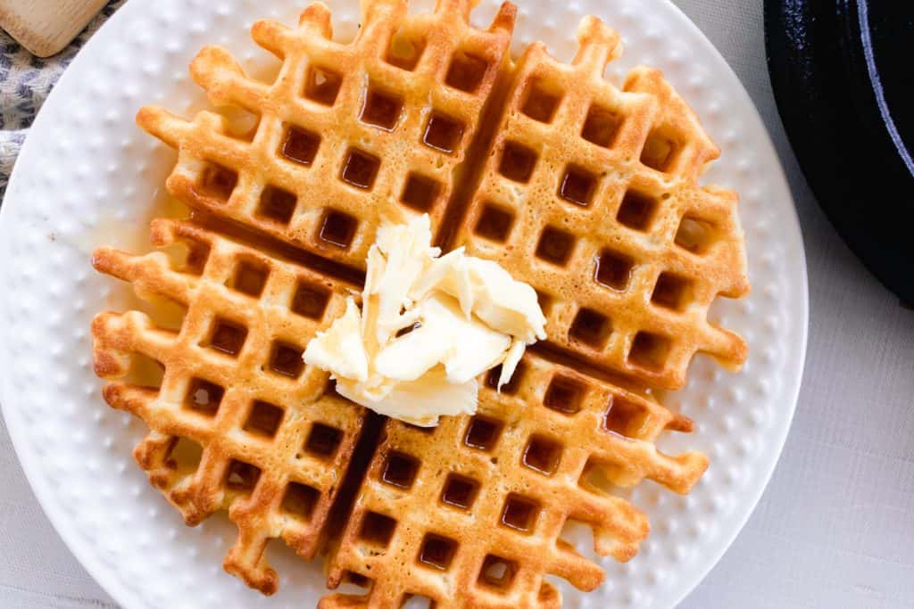 A golden waffle topped with butter and covered in maple syrup, served on a decorative white plate. 