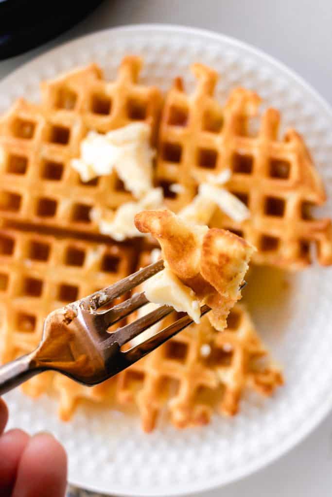 A fork with a serving of buttered golden waffle, with the rest of the waffle visible in the background on a decorative white plate.