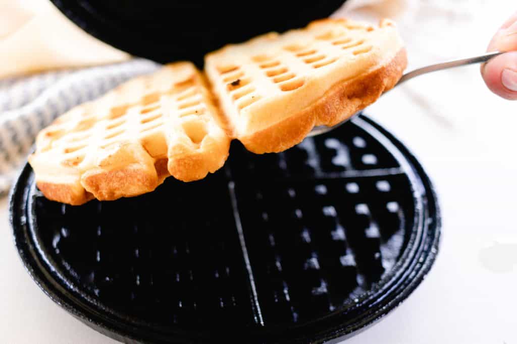 A golden waffle being lifted out of a black, cast iron waffle iron with a fork, beside a blue and white tea towel.