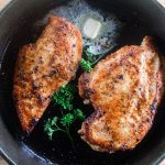 Chicken breasts searing in a cast iron skillet with a dollop of butter.