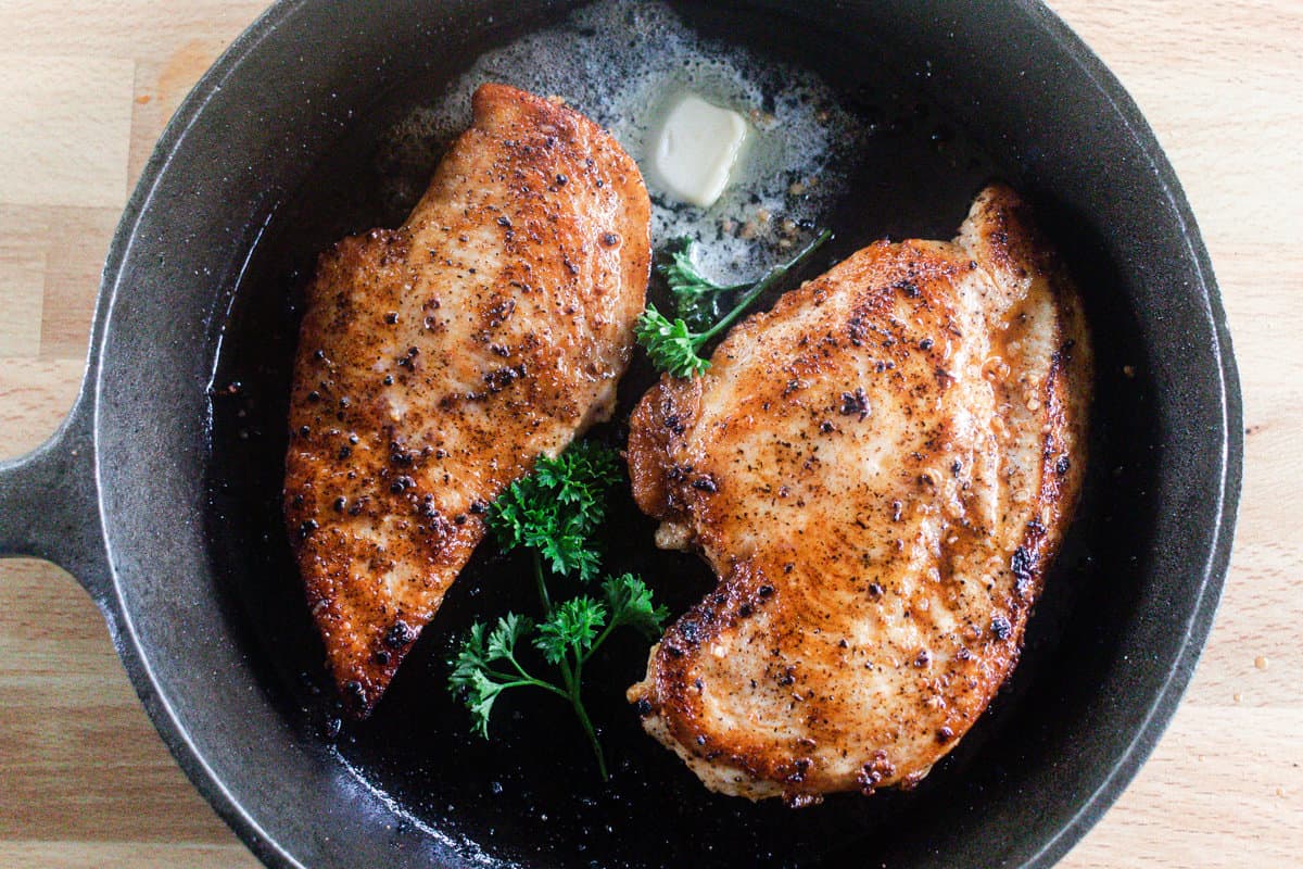 Chicken breasts searing in a cast iron skillet with a dollop of butter.