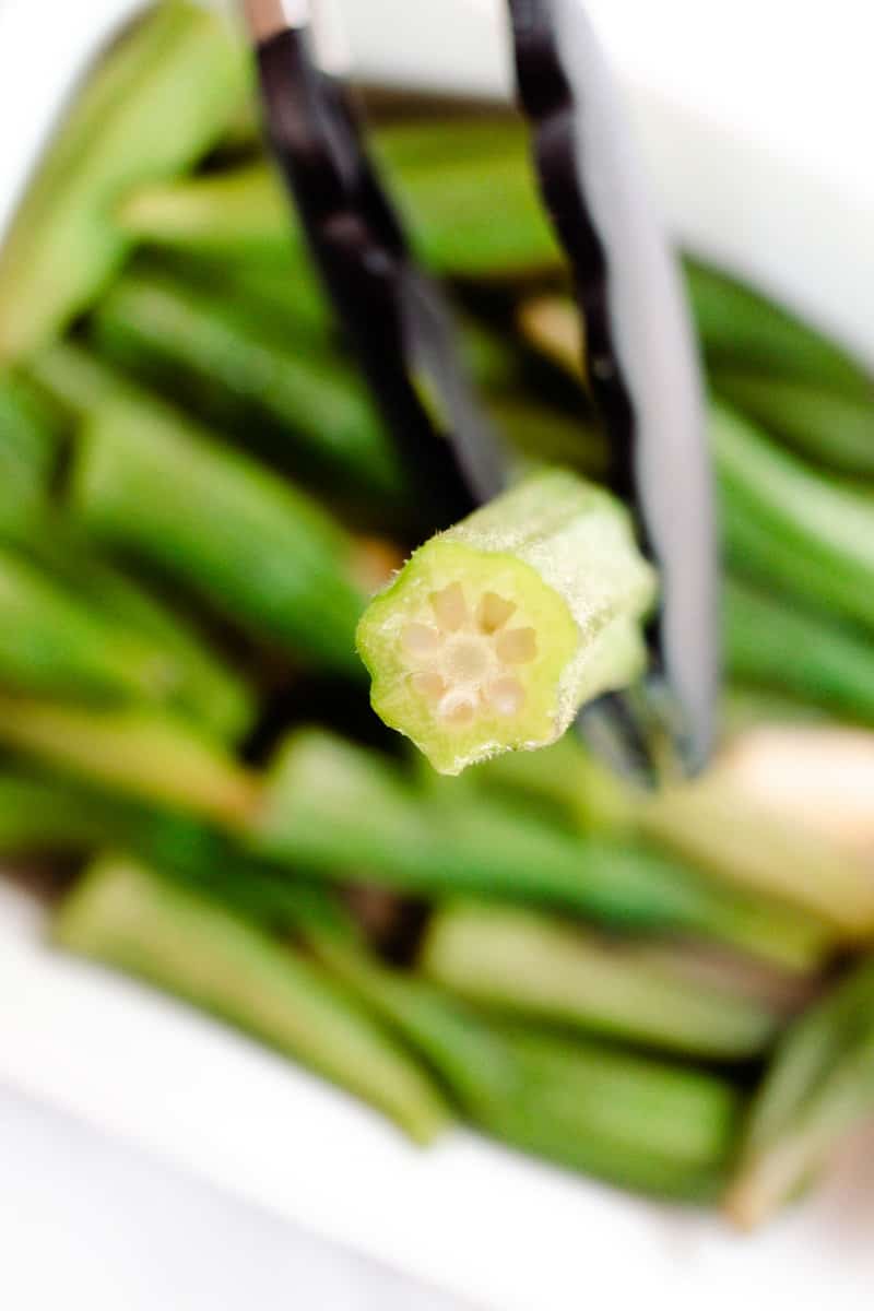 Focused on a tongs holding a piece of okra with a plate of okra in the background. 