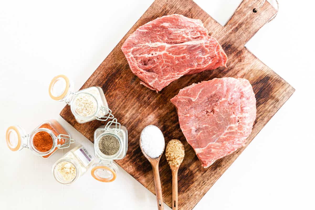 2 thick flat iron steaks with seasonings proportioned out on a wooden cutting board.